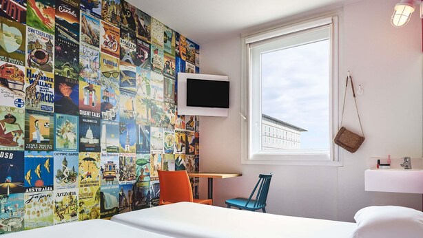 Et hotelværelse med en væg dækket af rejseplakater, et fjernsyn ophængt på væggen, et vindue med udsigt til en bygning, et lille bord, en stol og en seng med hvide sengetøj.