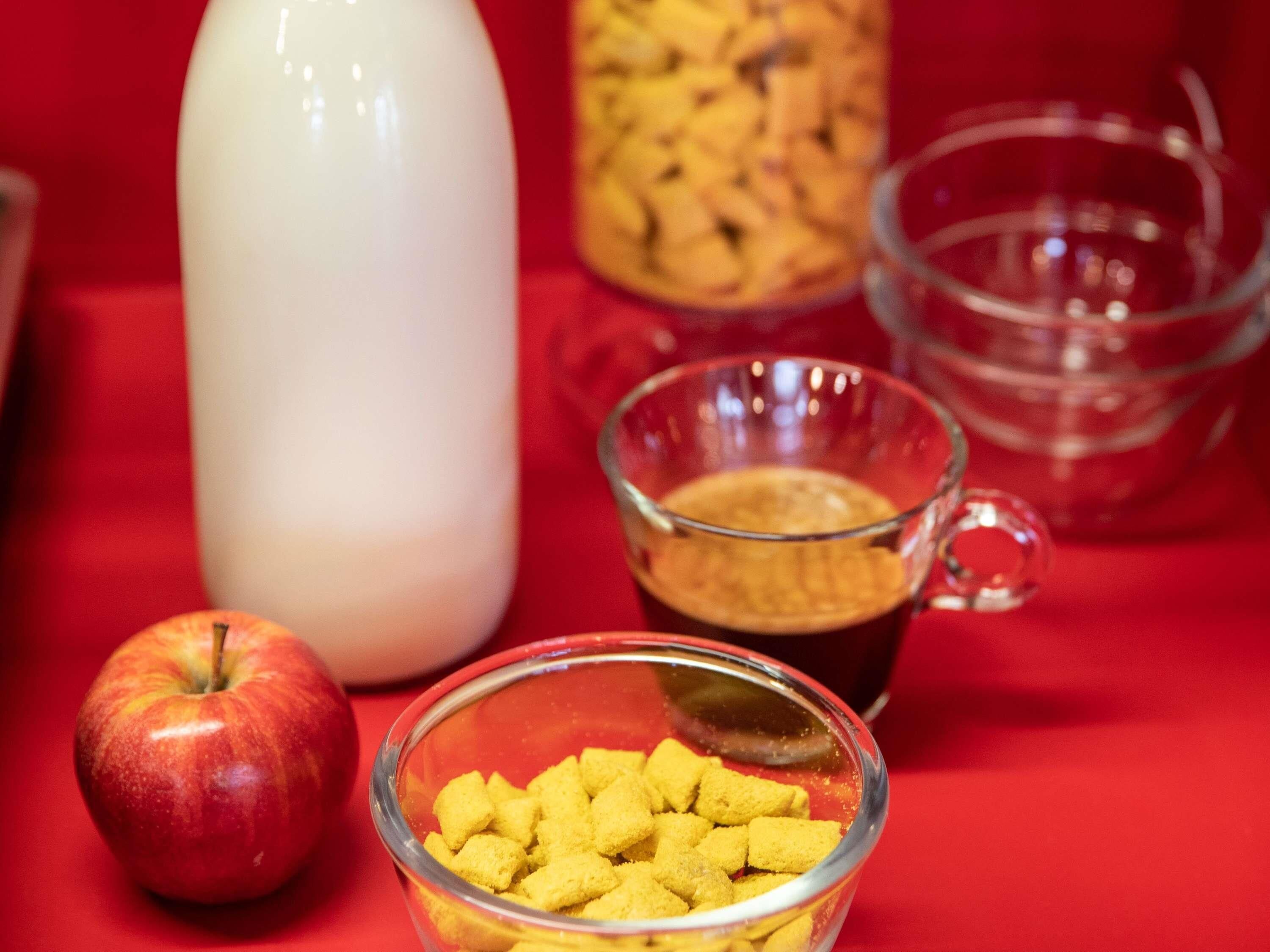A red surface with a glass bowl of yellow granules, a glass mug with a dark liquid, a red apple, and a white bottle.