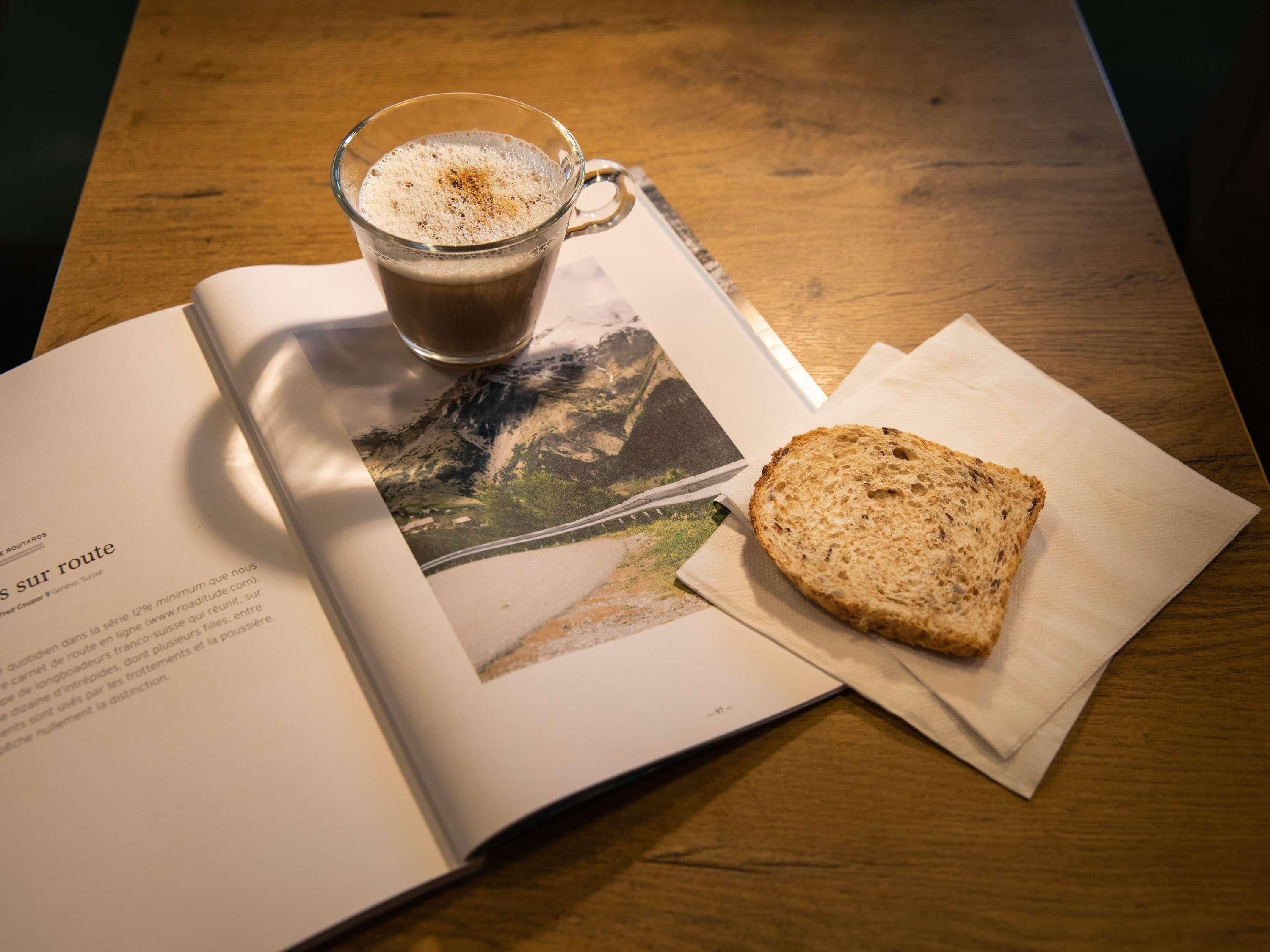 Un livre, une tasse de café et une tranche de pain sont posés sur une table, et une photographie se trouve à proximité.