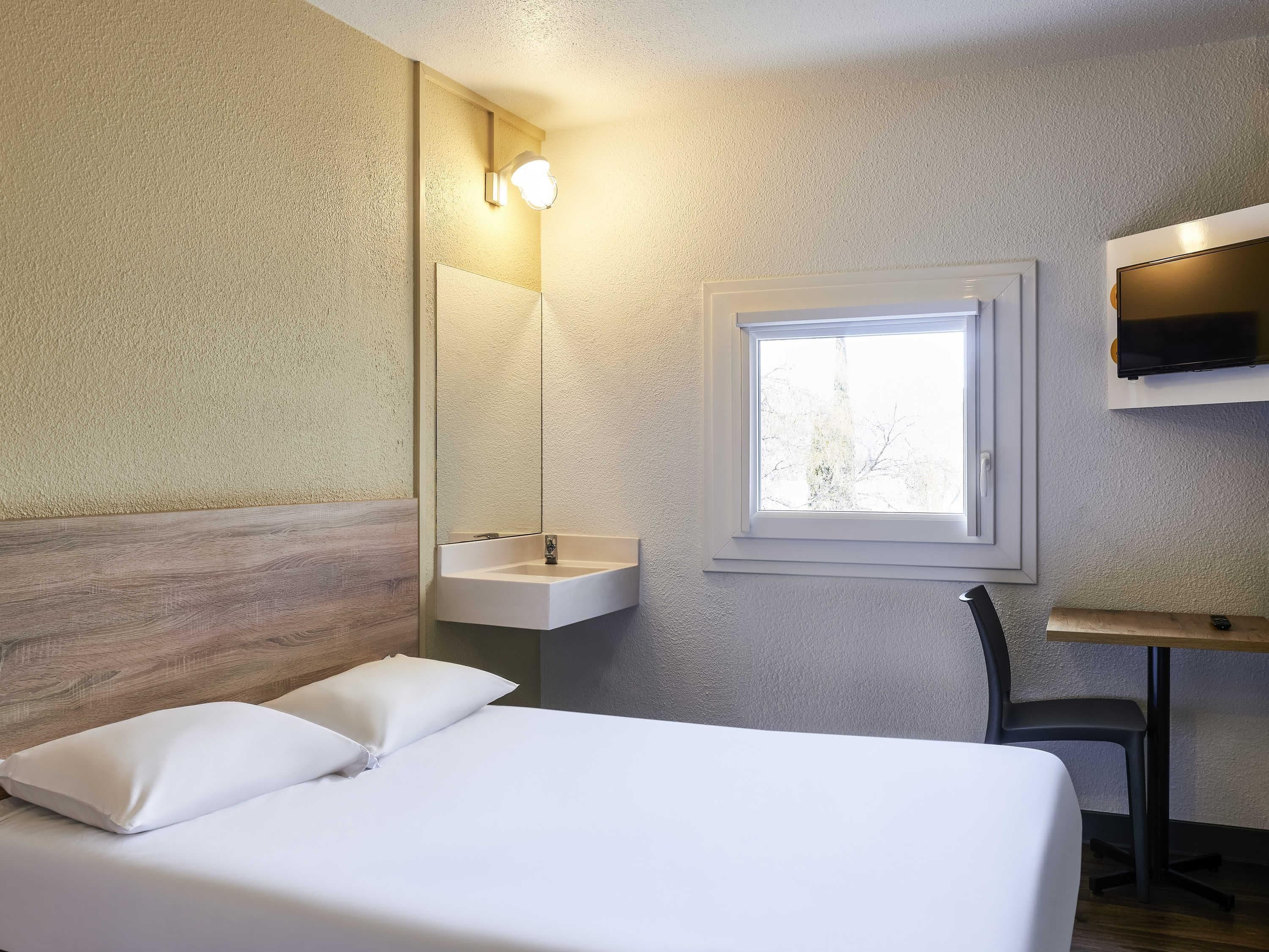 Un dormitorio con cama, mesita de noche, espejo, ventana y televisor instalado en la pared.