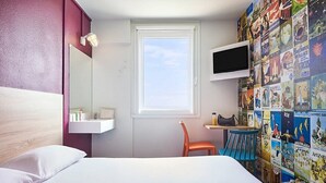 A hotel room with a bed, a chair, a small table, a TV mounted on the wall, and a window with a view of the sky. - hotelF1 Beauvais (Beauvais)