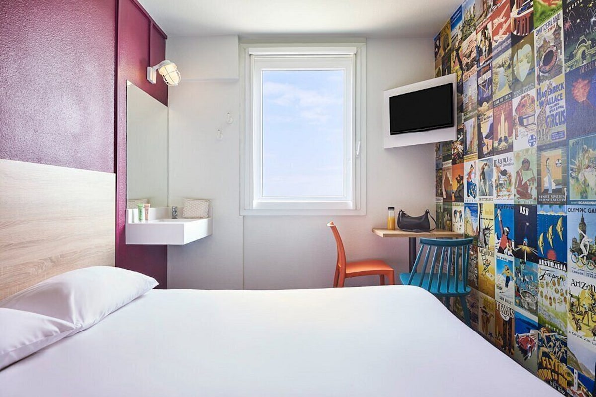 A hotel room with a bed, a chair, a small table, a TV mounted on the wall, and a window with a view of the sky.