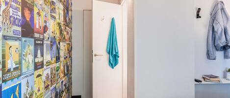 A hotel room with a bed, a door with a towel hanging, a wall with travel posters, and a small desk with a book and a phone.