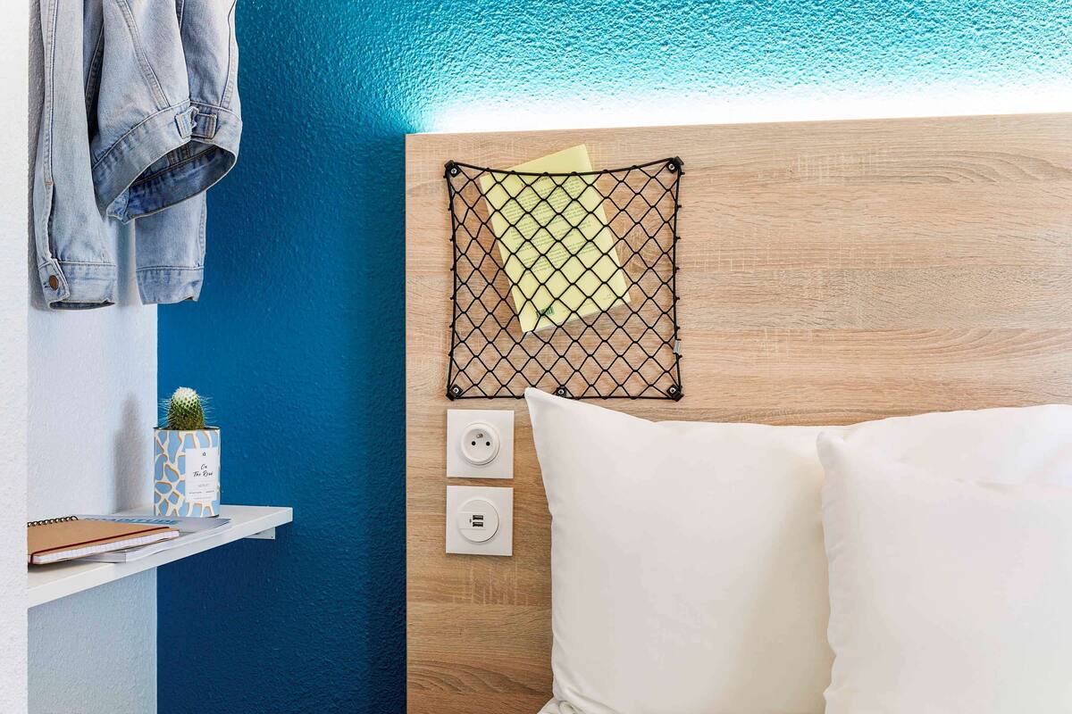 A neatly arranged hotel room with a wooden headboard, a bed with white linens, a small shelf with a cactus and a notebook, and a wall-mounted power outlet.