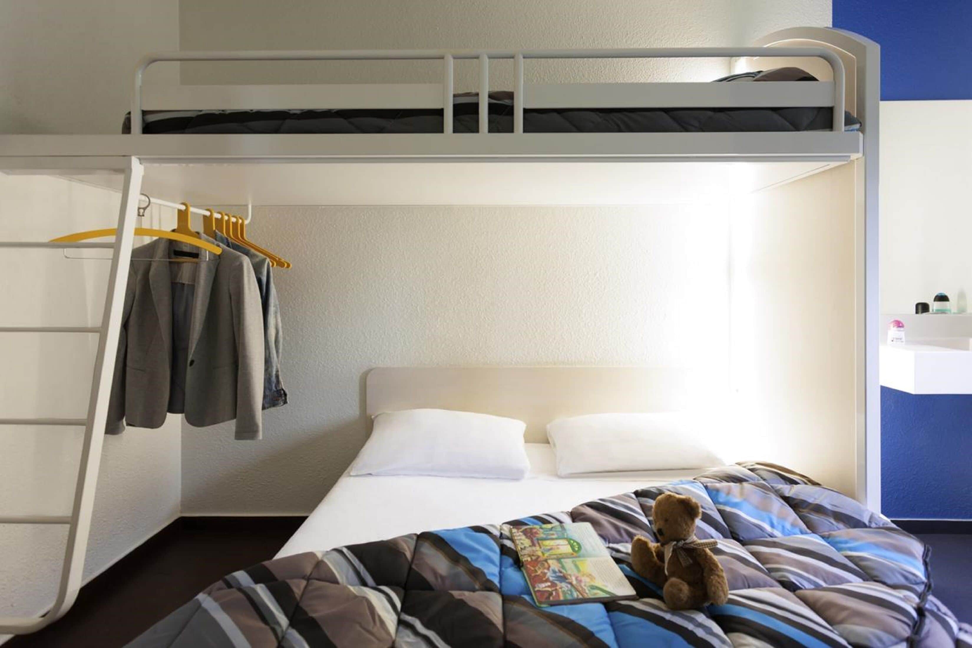 Habitación con literas, cabecero blanco, perchero con algunas prendas, un osito de peluche en la cama inferior y un pequeño estante con artículos de aseo.
