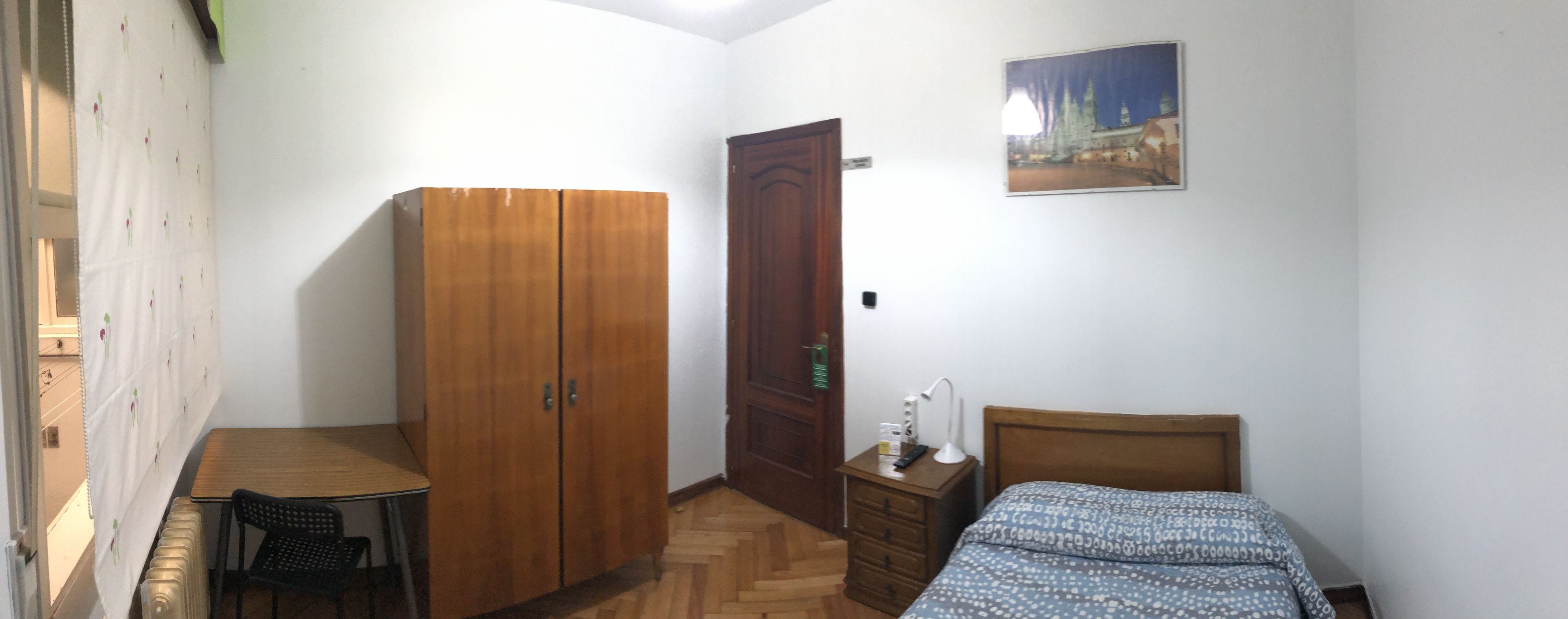 Un dormitorio con un armario de madera, una cama con ropa de cama estampada, una mesita y una silla.
