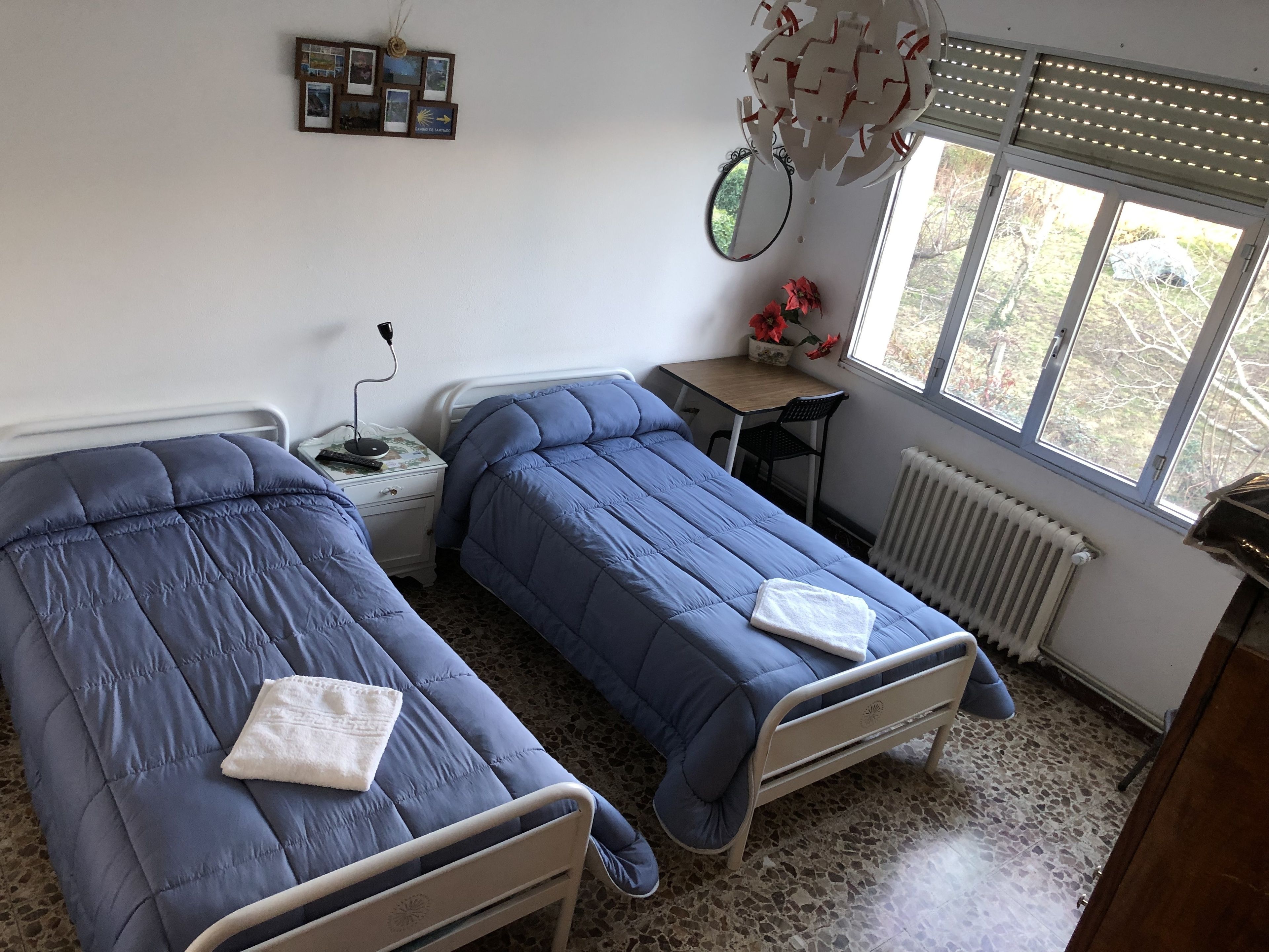 Ein Zimmer mit zwei Betten, einem kleinen Tisch, einem Stuhl, einem Heizkörper und einem Fenster mit Blick auf Bäume.