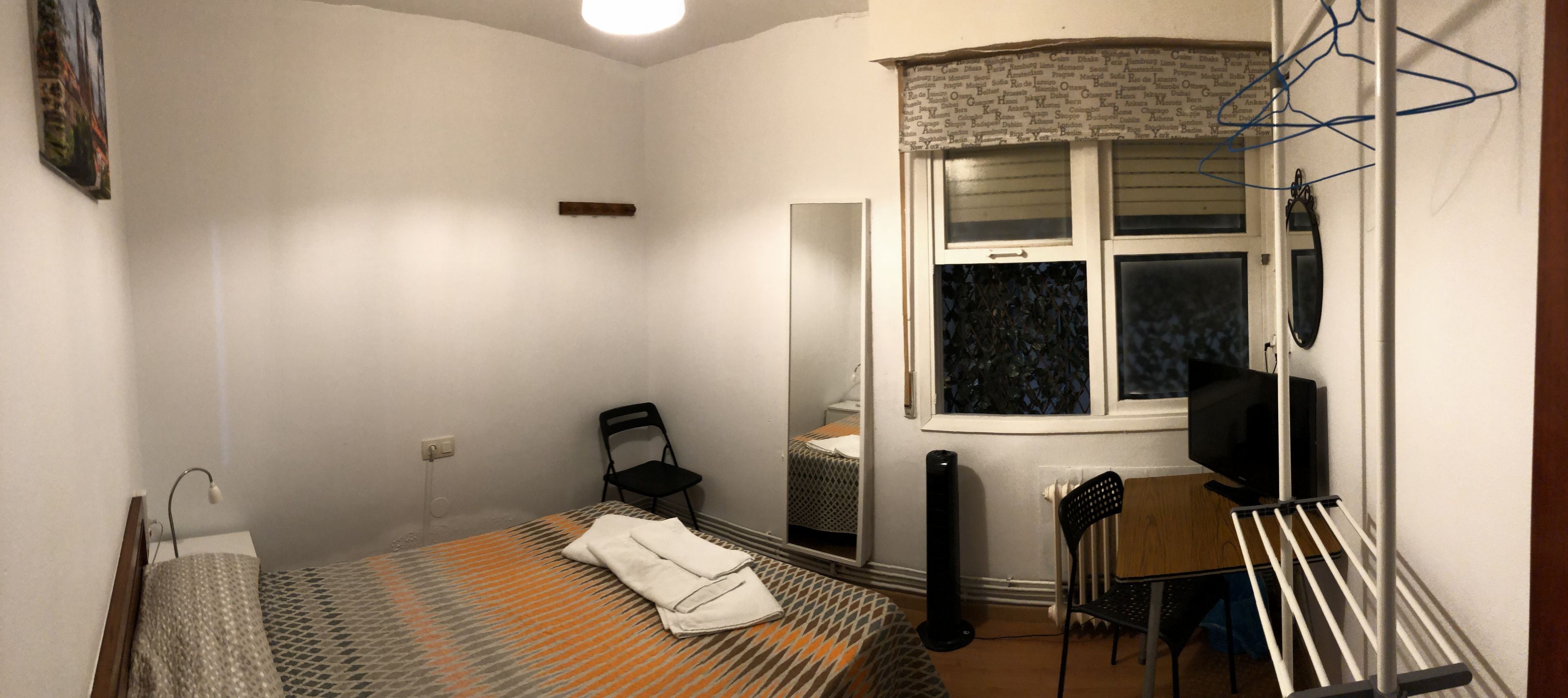 Una habitación pequeña y poco amueblada con una cama individual, una silla, un escritorio, un espejo y una ventana con persianas.