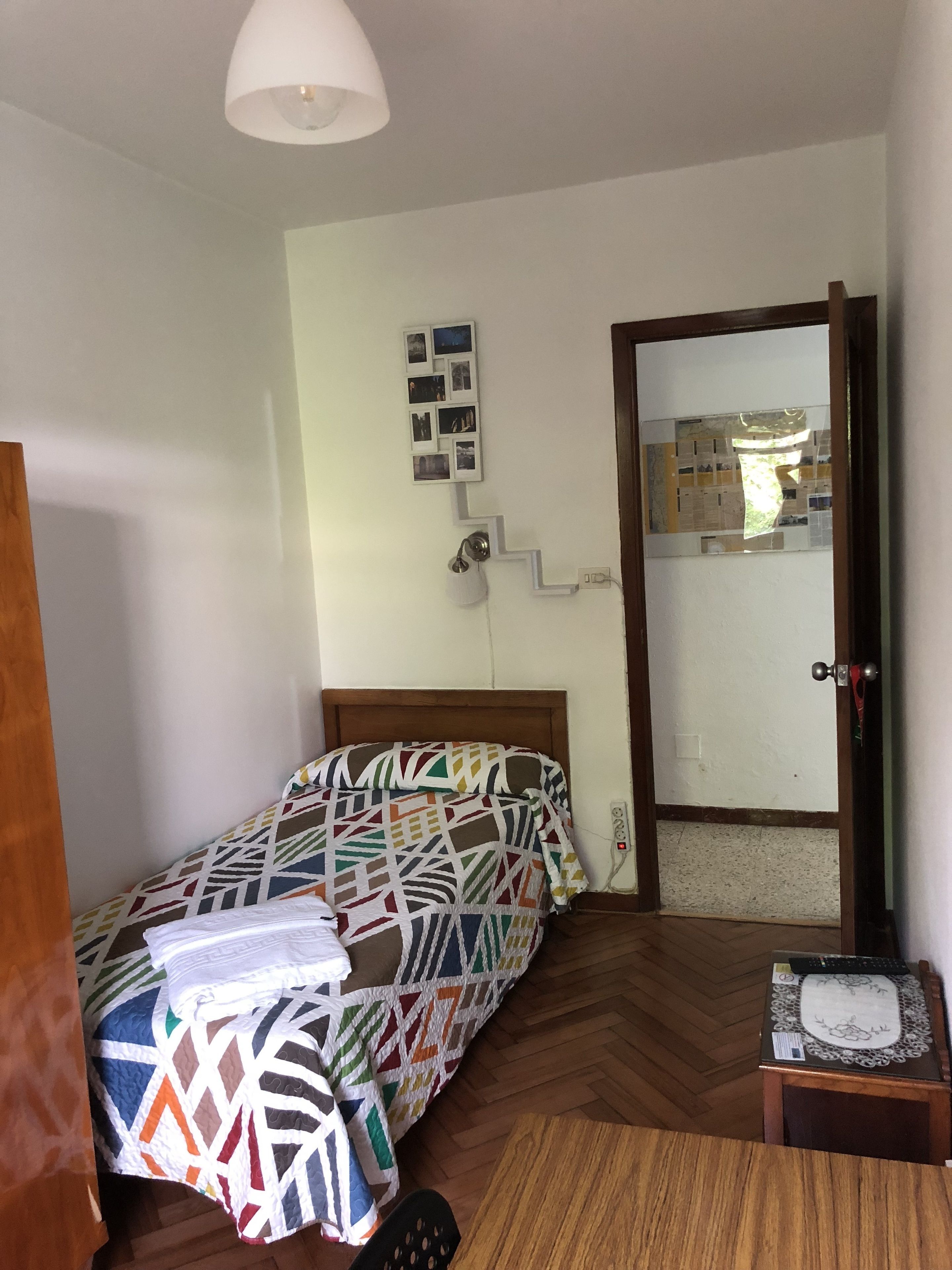 Un dormitorio pequeño y ordenado con cama, mesita de noche, espejo y repisa con fotografías.