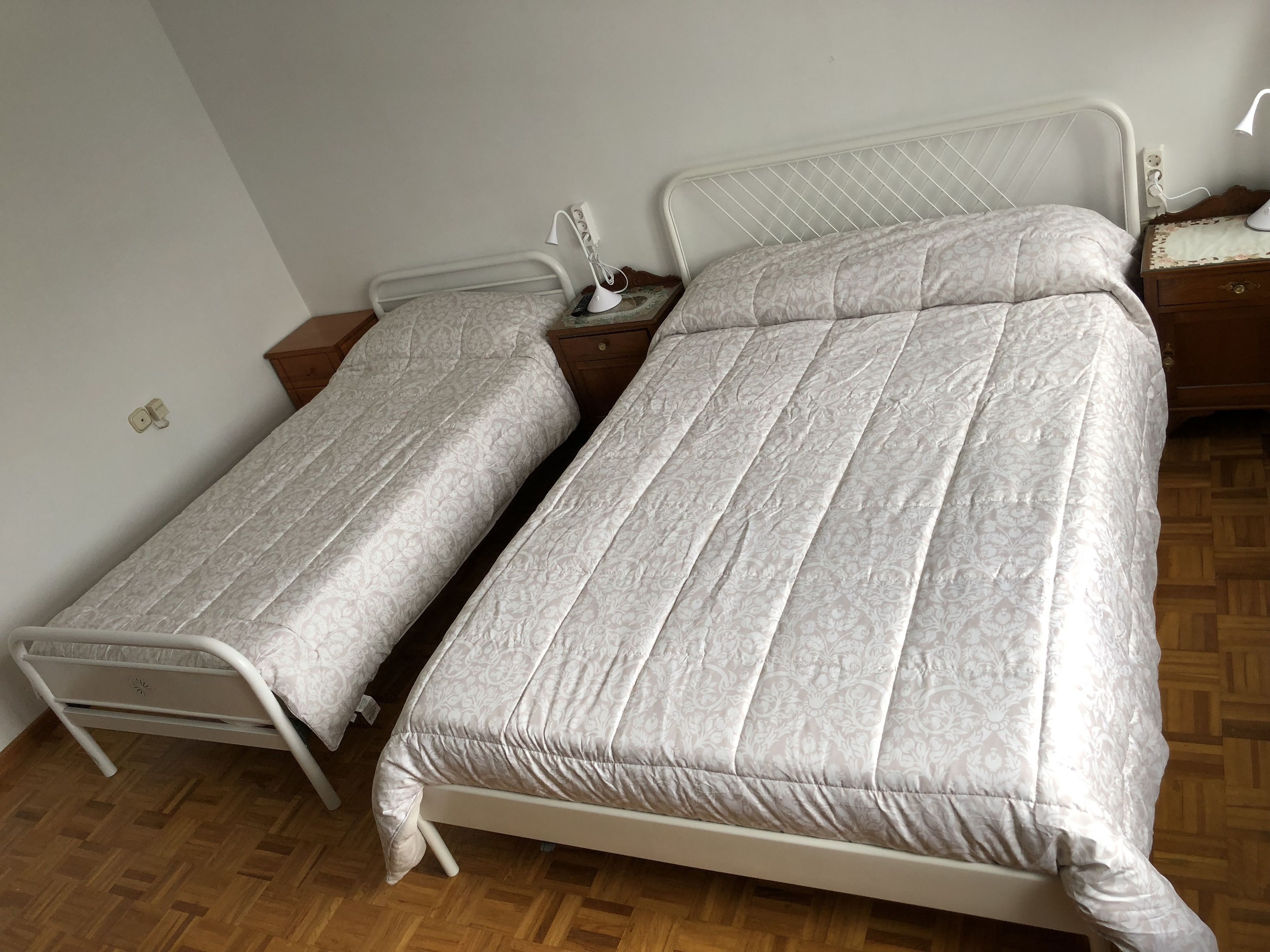 Een tweepersoonsbed met een wit metalen frame en bijpassend hoofdbord, een nachtkastje met een lamp en een houten nachtkastje.