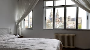 A bedroom with a large bed, a nightstand, a lamp, a radiator, and a view of a cityscape through a window. - Hostal Costa Azul (Santiago de Compostela)