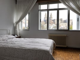 Un dormitorio con una cama grande, una mesita de noche, una lámpara, un radiador y vistas a un paisaje urbano a través de una ventana.