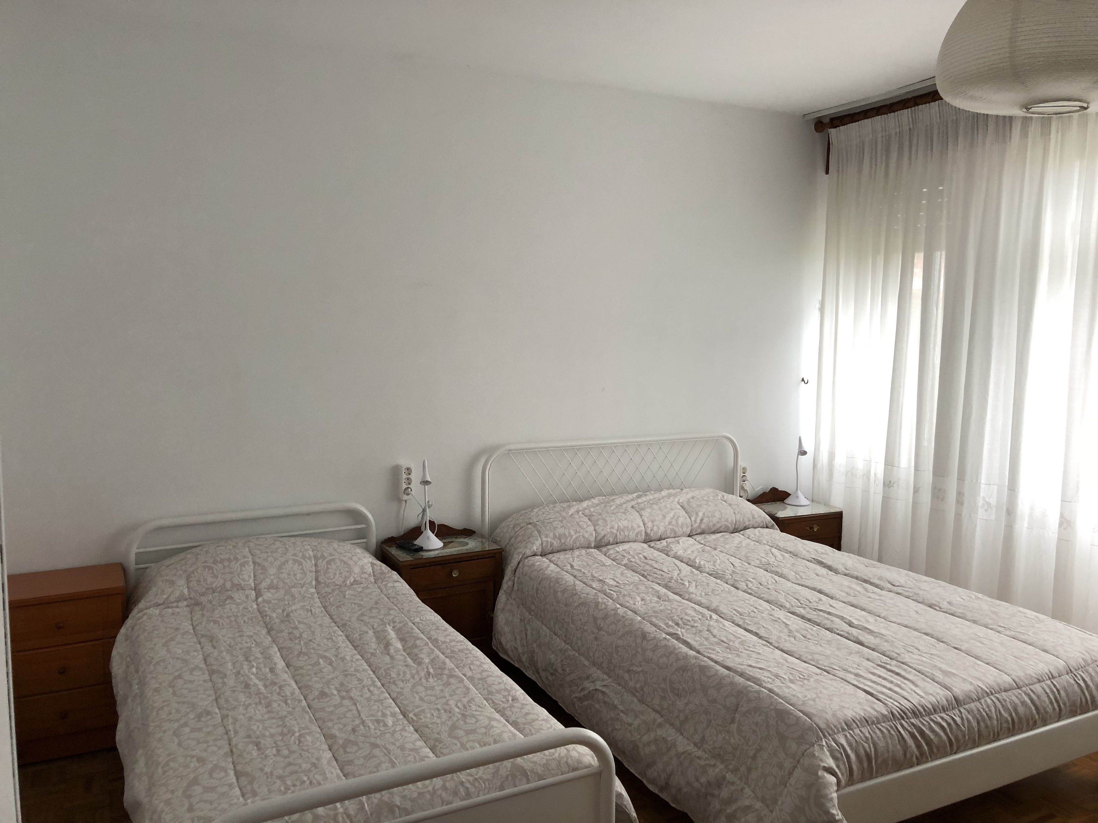 Un dormitorio con dos camas individuales, una mesita de noche con lámpara y una ventana con cortinas translúcidas.