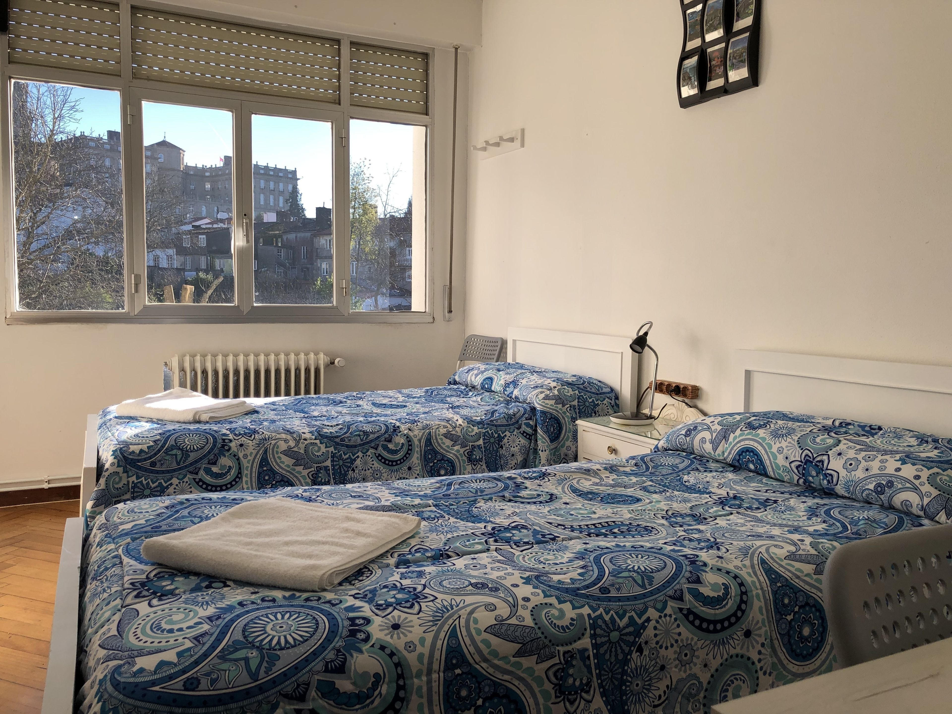 A bedroom with two beds, a radiator, a window with blinds, and a view of buildings outside.