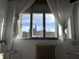 Una habitación con un ventanal grande, cortinas blancas, un radiador y dos sillones.