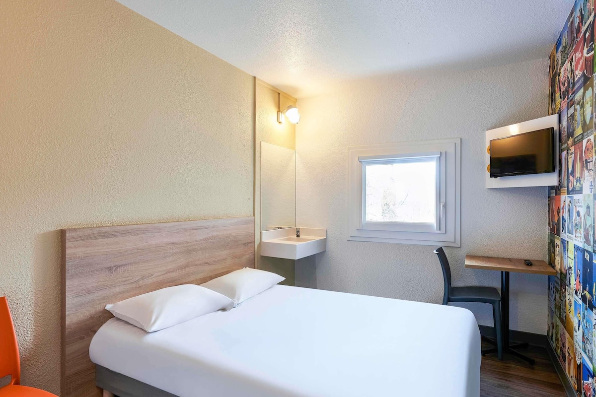 A hotel room with a bed, a desk, a chair, a TV, a window, and a wall with a poster.