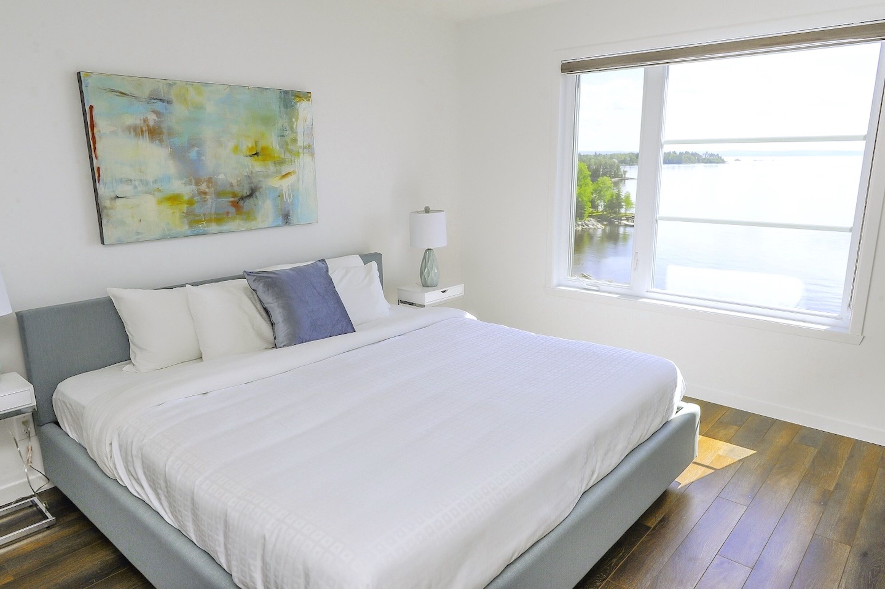 A modern bedroom with a large bed, a nightstand, a lamp, and a window with a view of a lake.