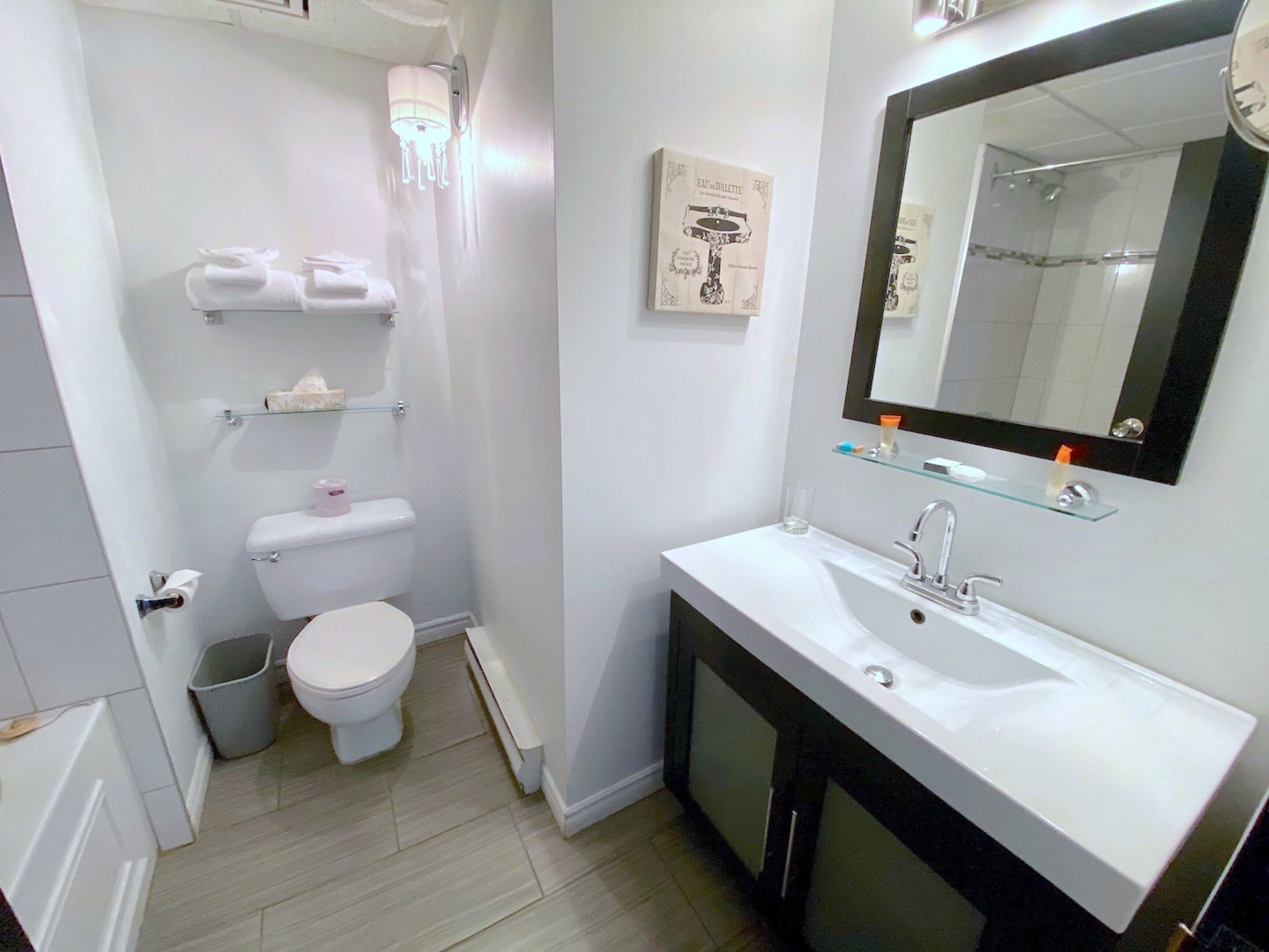 A modern bathroom with a white sink, a mirror, and a toilet. There are shelves with toiletries and a trash bin.
