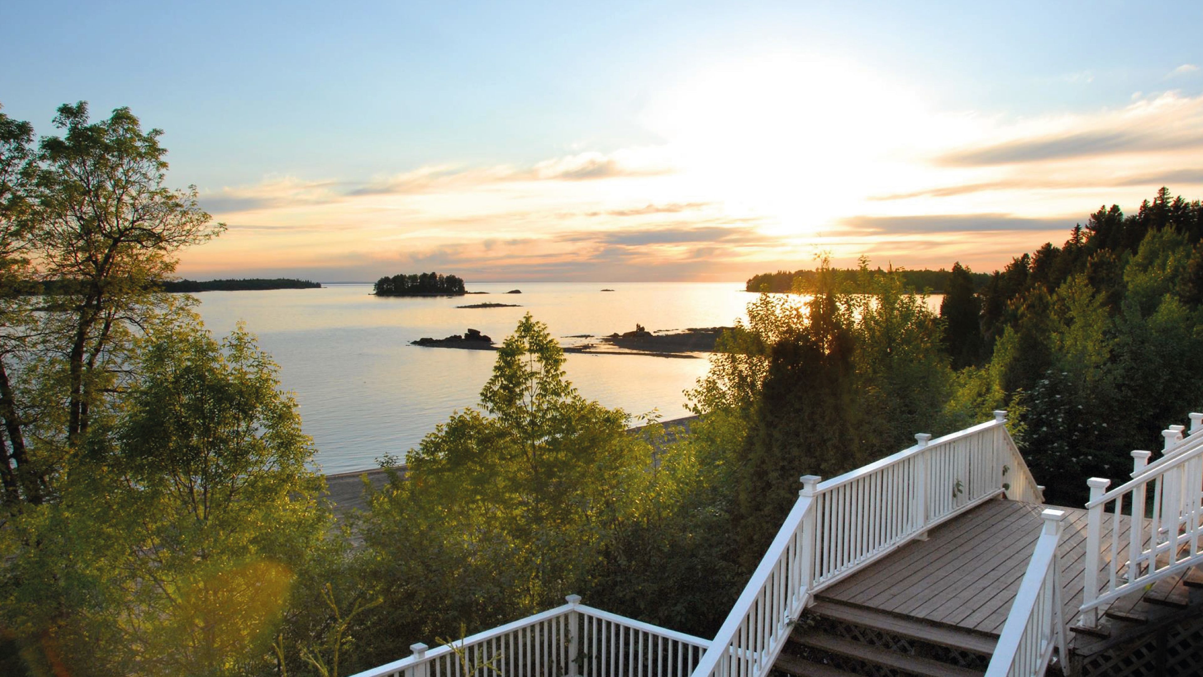 A wooden deck with white railings overlooking a calm lake with distant islands and a sunset in the background.