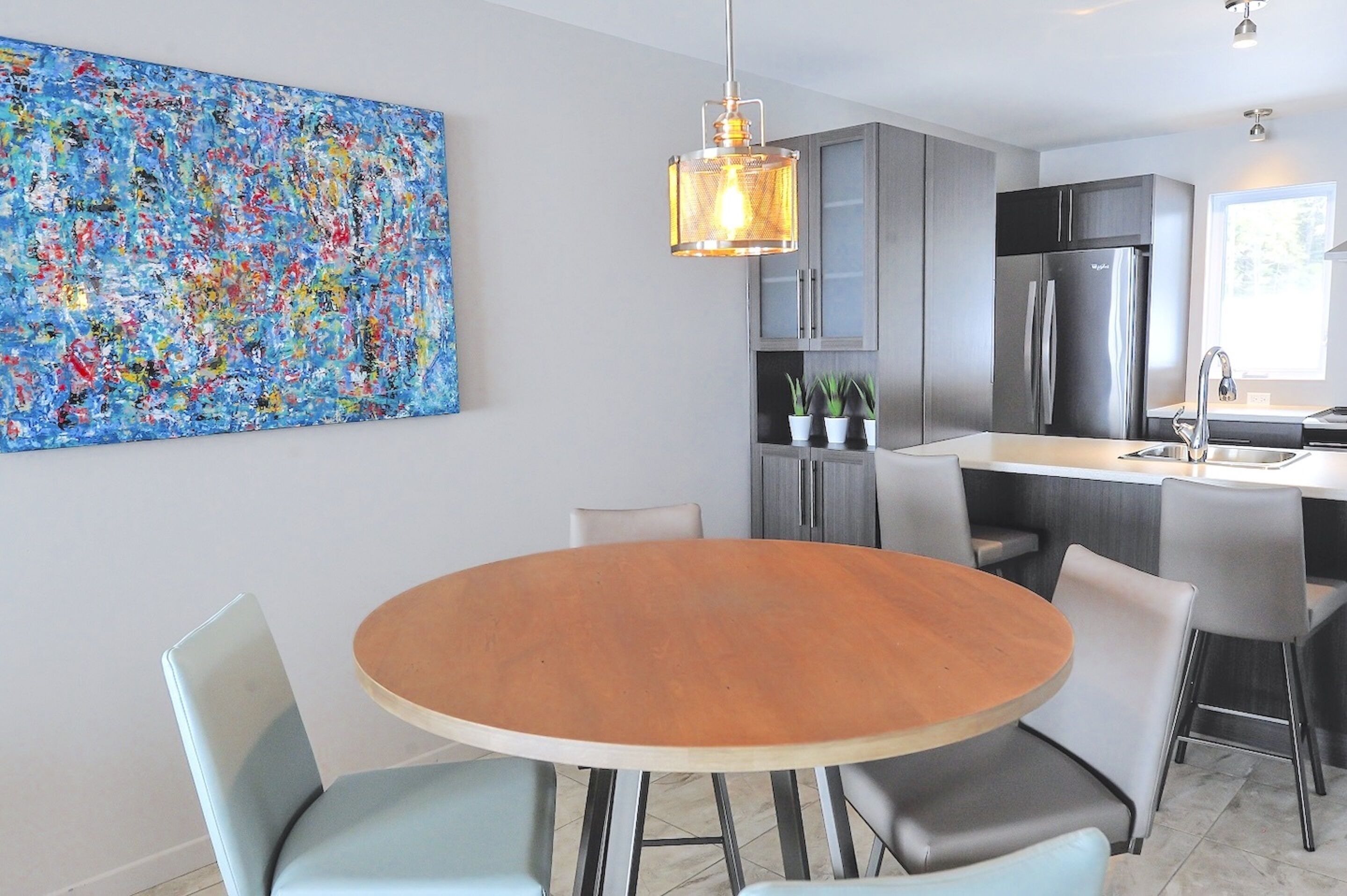 A modern kitchen with a stainless steel refrigerator, a round wooden table, and a hanging pendant light.