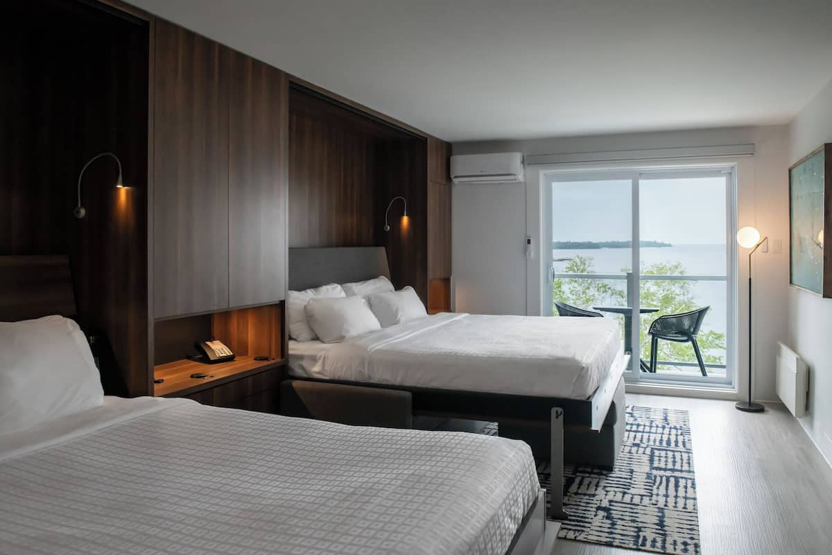 Ein Hotelzimmer mit zwei Betten, einem Holzschrank, einem Nachttisch mit Lampe, einem kleinen Tisch mit Stuhl und einem großen Fenster mit Meerblick.