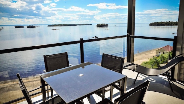 A balcony with a table and chairs overlooking calm water and distant islands. - Auberge des Îles (Saint-Gedeon)