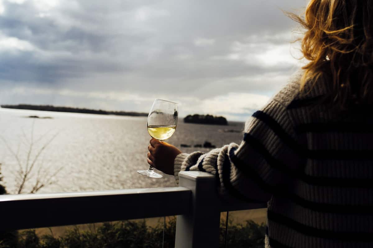Eine Person hält ein Glas Weißwein und blickt auf ein Gewässer mit bewölktem Himmel.