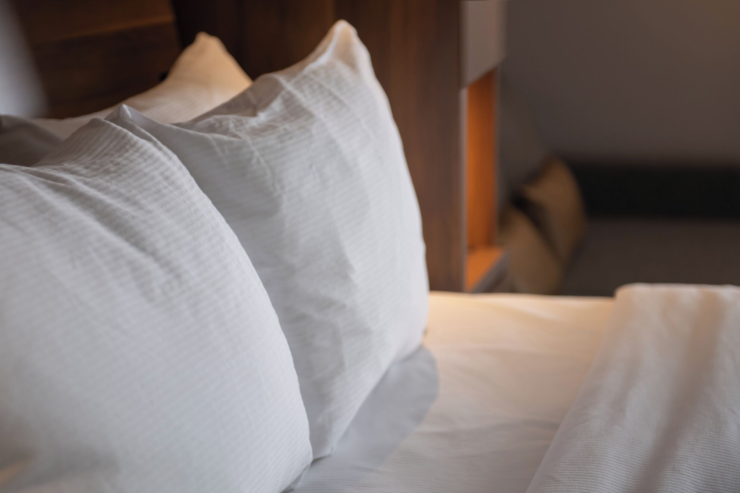 Un lit avec du linge de lit blanc et des oreillers, une tête de lit en bois et un mur de couleur neutre en arrière-plan.