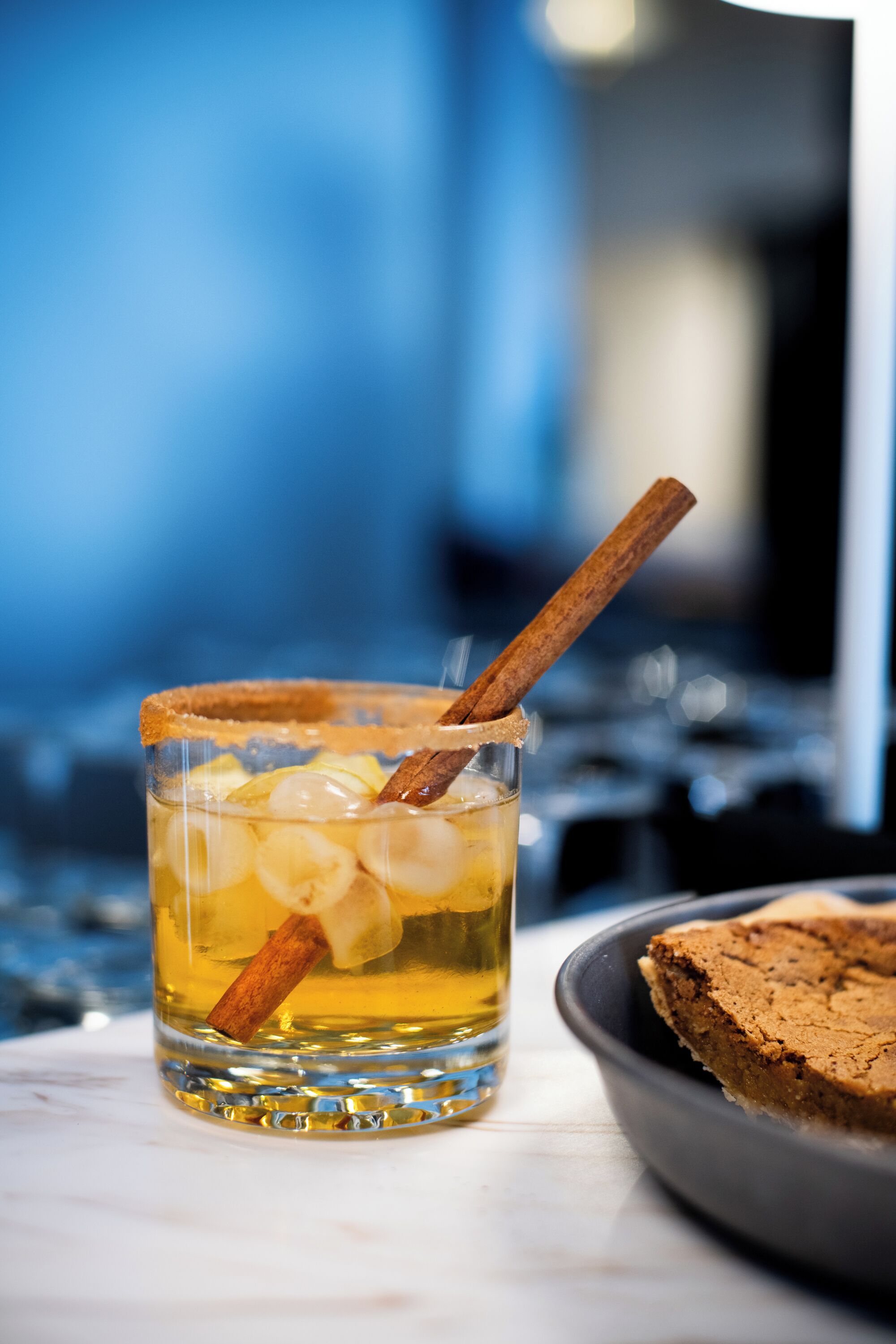 Un verre d’un liquide ambré, agrémenté d’une tige de cannelle, accompagné d’une part de gâteau présentée sur une assiette.