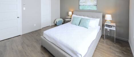 A bedroom with a large bed, two bedside tables with lamps, a small chair, and a painting on the wall.
