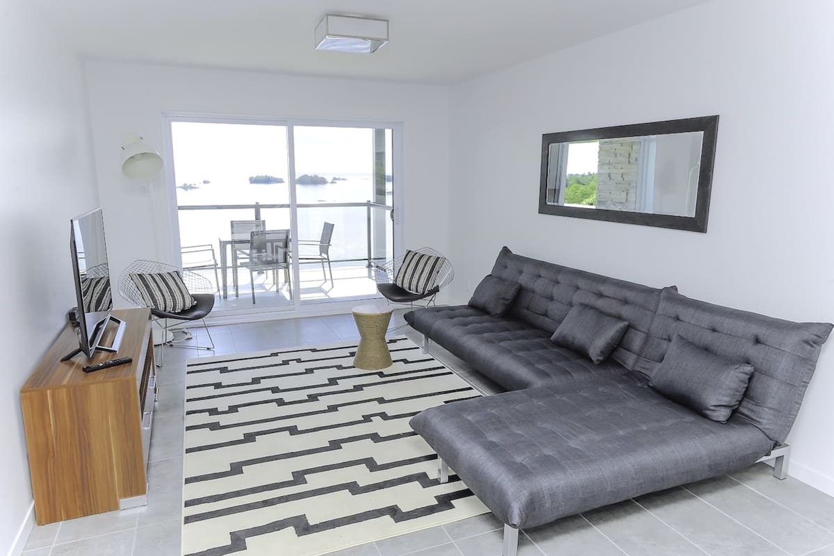 Ein modernes Wohnzimmer mit einer grauen Eckcouch, einem Holztisch, einem Fernseher und einem großen Fenster mit Blick aufs Meer.