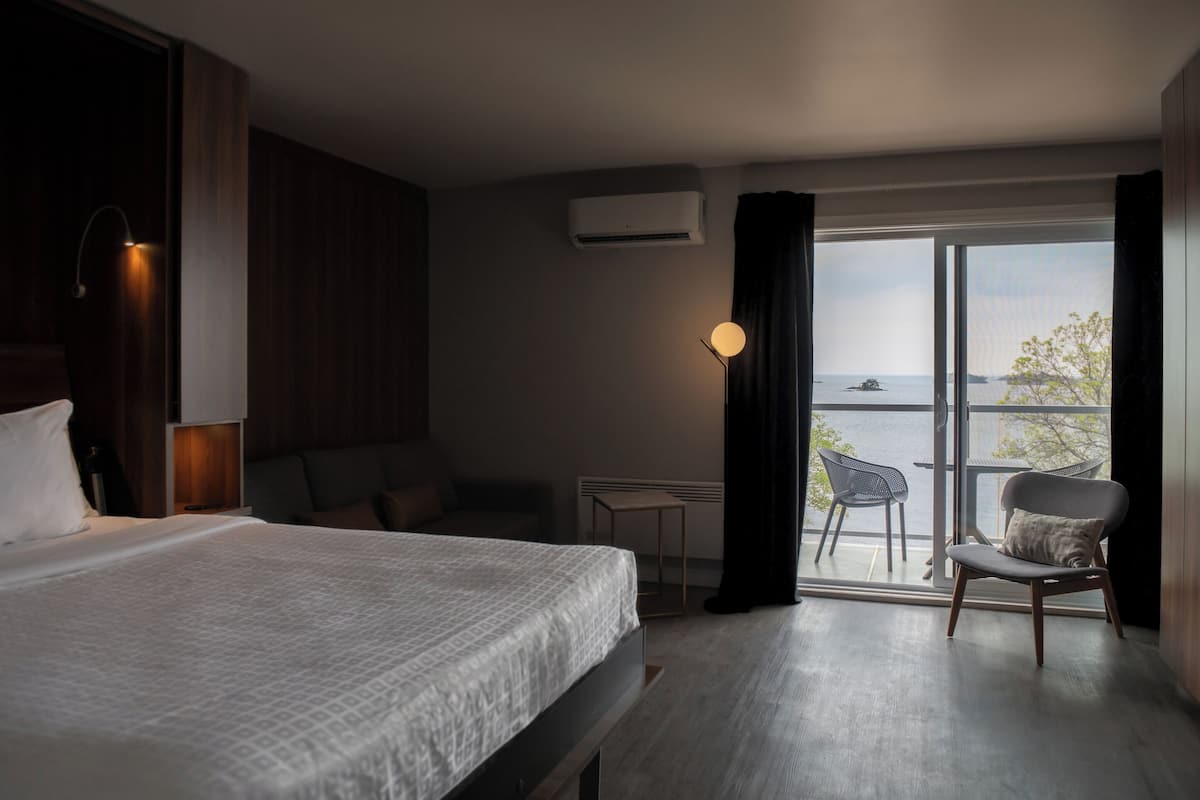 Ein modernes Hotelzimmer mit einem großen Bett, einem kleinen Tisch, einem Sessel und Meerblick durch ein großes Fenster.