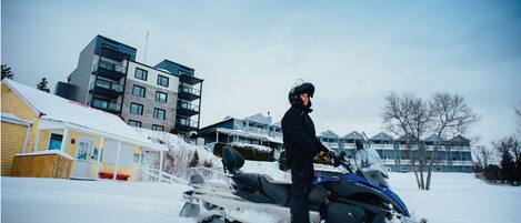 A person on a snowmobile in a residential area with multi-story buildings in the background.