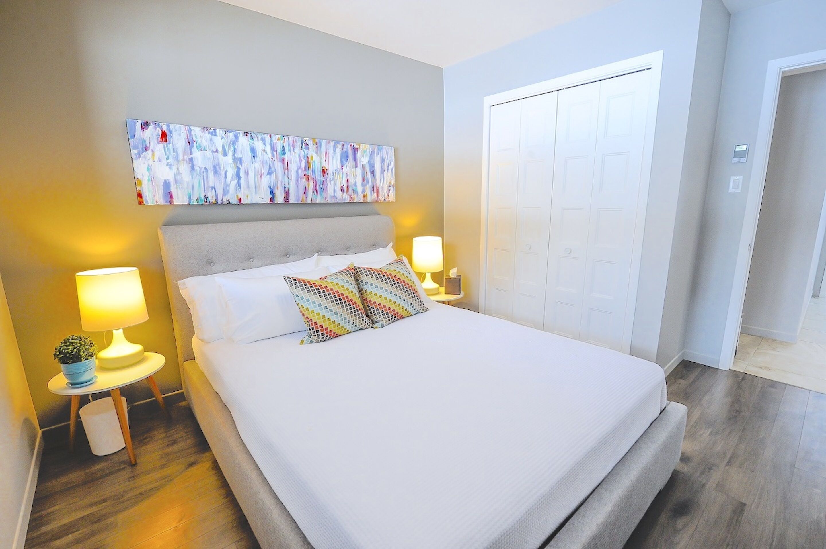 Un dormitorio ordenado con cama, dos lámparas de noche, una mesita con una planta y un cuadro abstracto de colores en la pared.