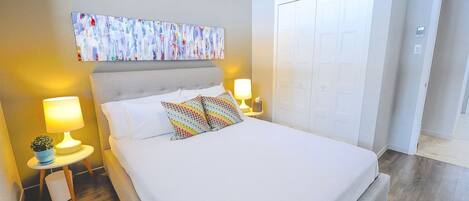 Un dormitorio ordenado con cama, dos lámparas de noche, una mesita con una planta y un cuadro abstracto de colores en la pared.