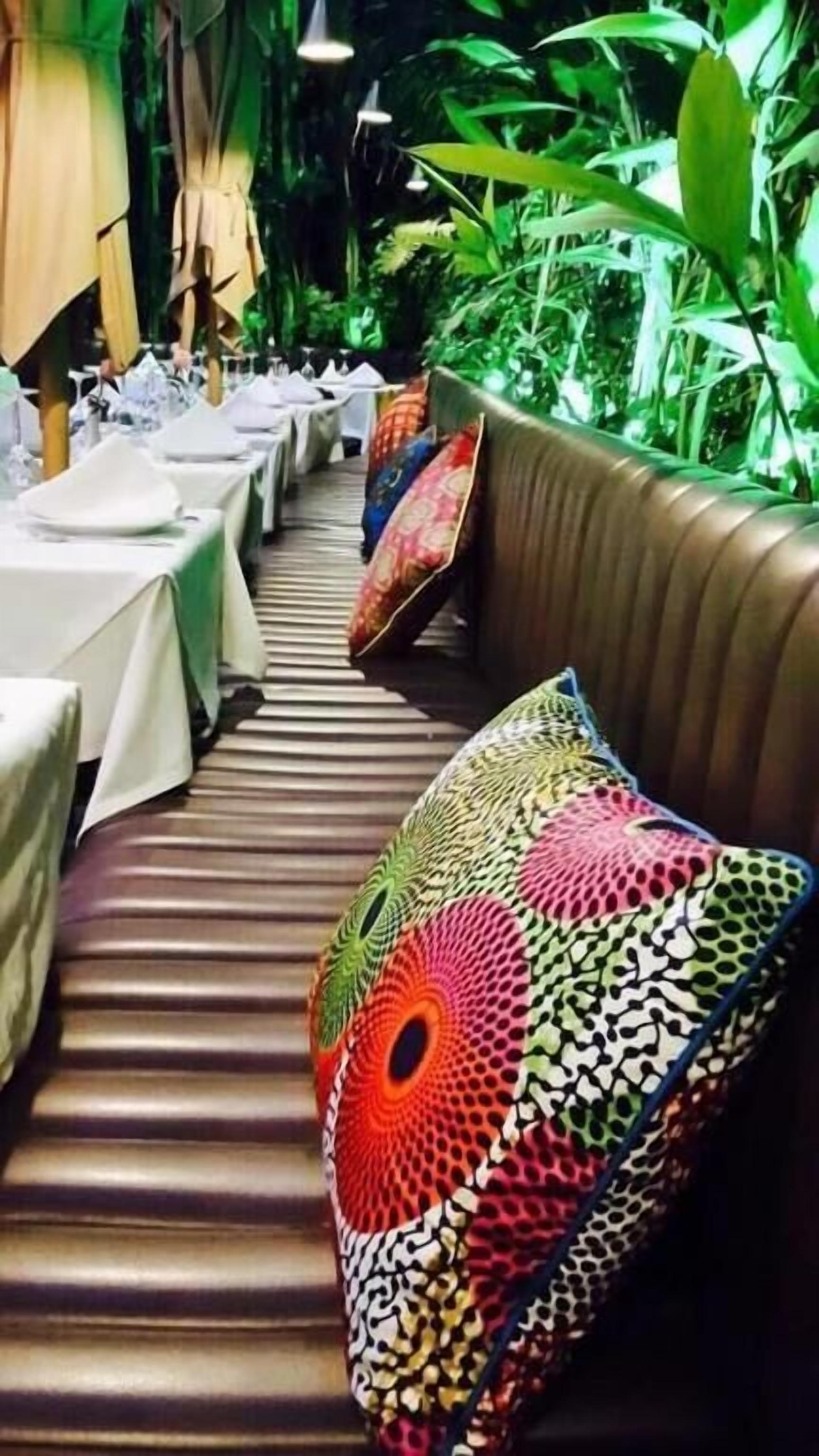 A restaurant with long tables covered in white tablecloths, cushioned booths with colorful patterned cushions, and a backdrop of green foliage.