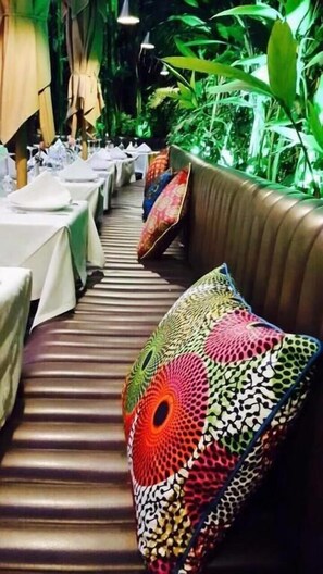 A restaurant with long tables covered in white tablecloths, cushioned booths with colorful patterned cushions, and a backdrop of green foliage.