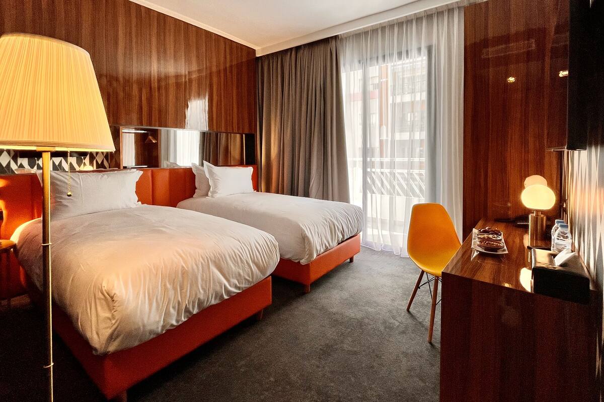 A hotel room with two beds, a desk, a chair, a lamp, and a TV.