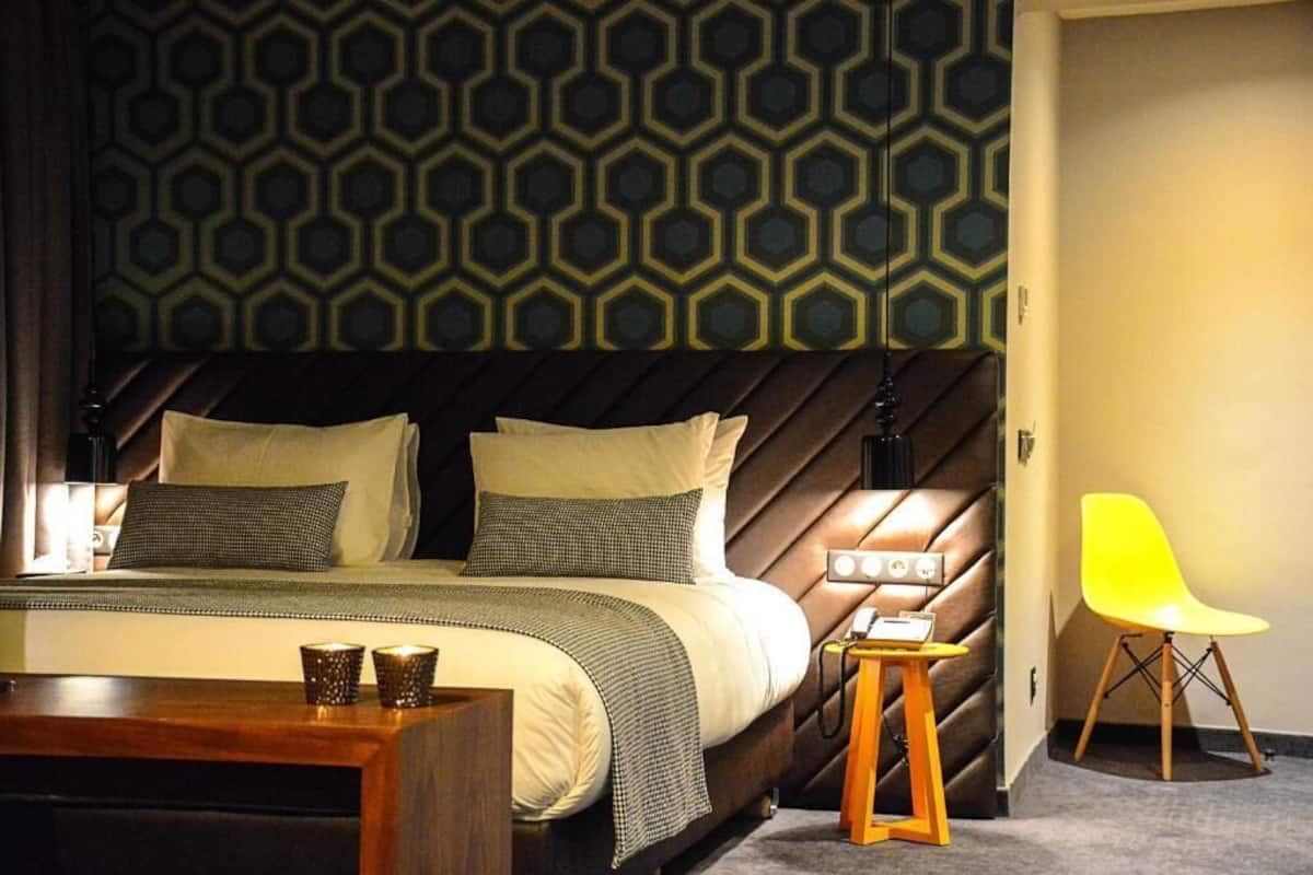 A modern hotel room with a large bed, a headboard with geometric patterns, a bright yellow chair, and a small wooden side table.