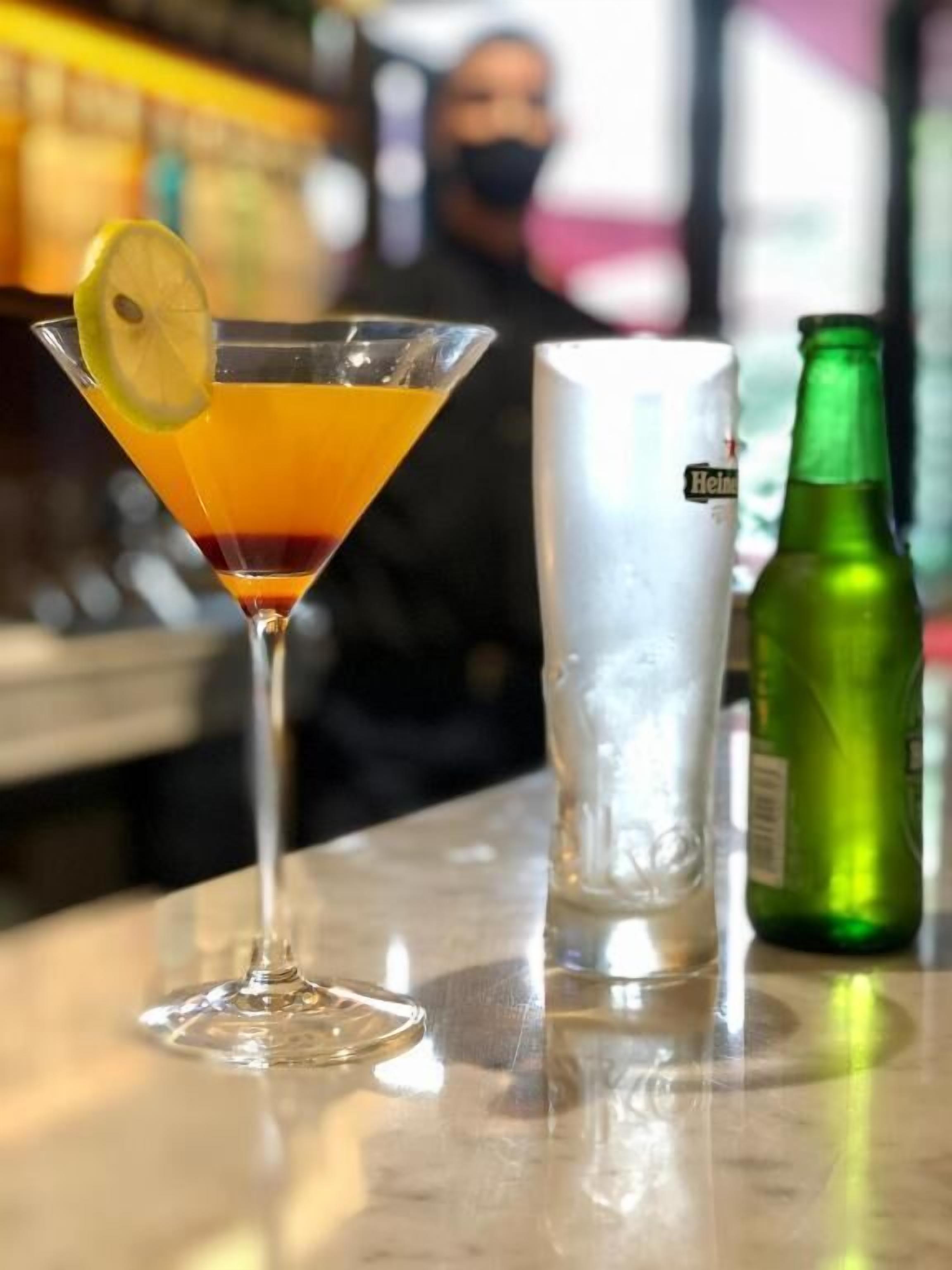 A martini glass with a lemon slice on a bar counter, a bottle of Heineken beer, and a large ice bucket.