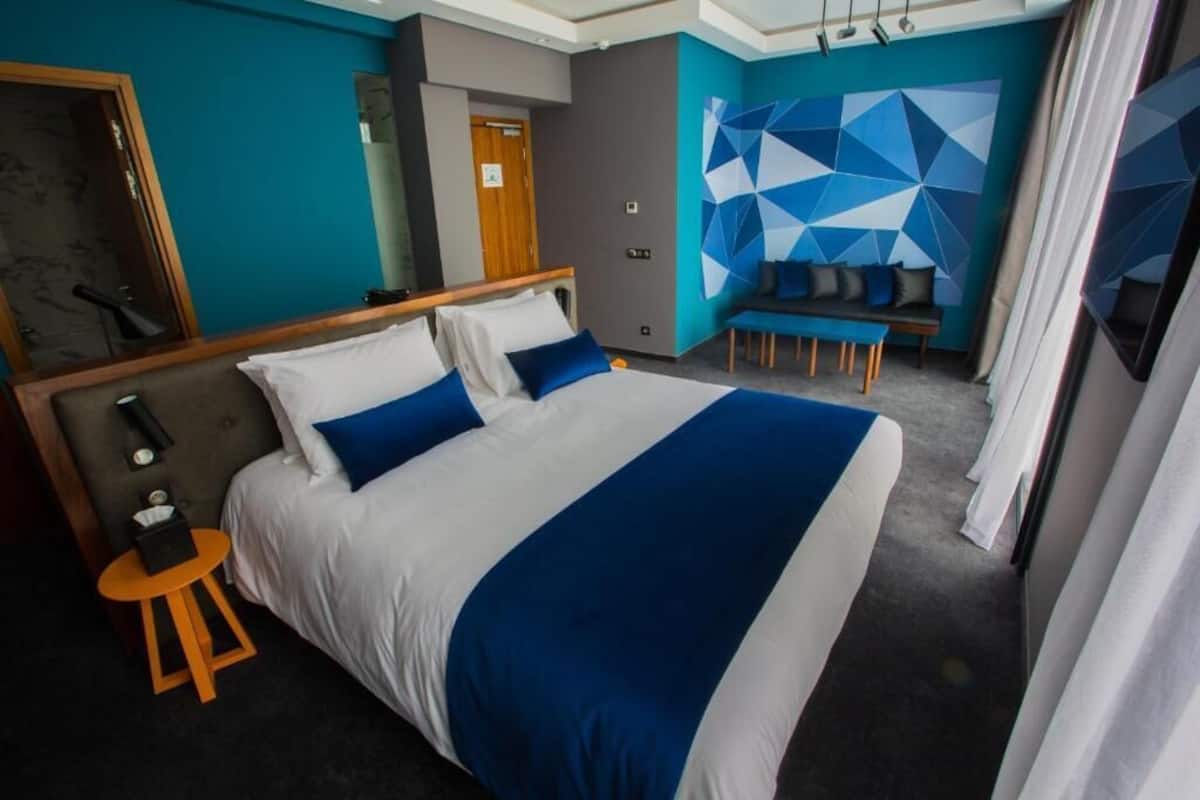 A modern hotel room with a large bed, a seating area with a sofa and chairs, a small table, and a mirror.