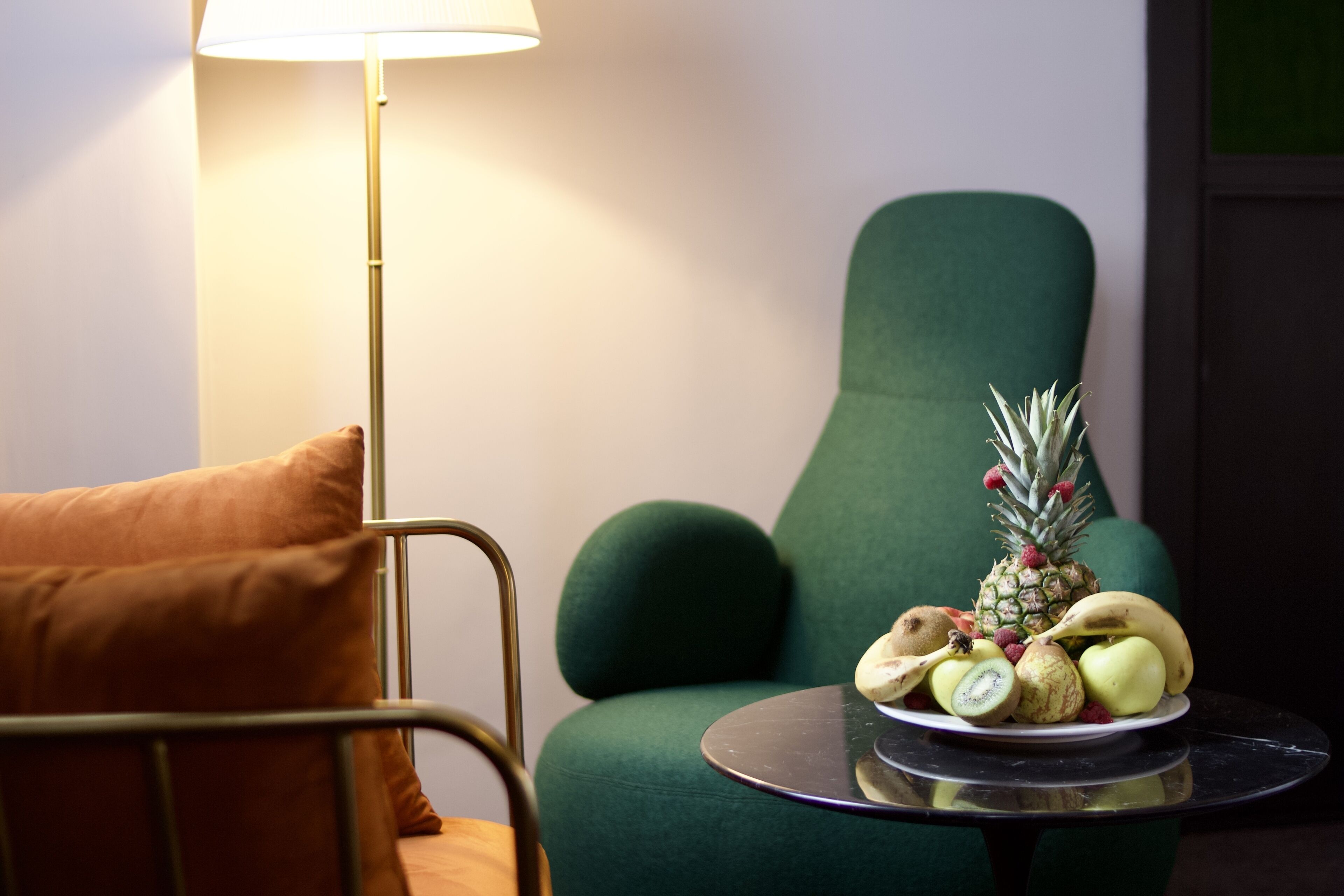 A green armchair, a small round table with a fruit plate, and a floor lamp.