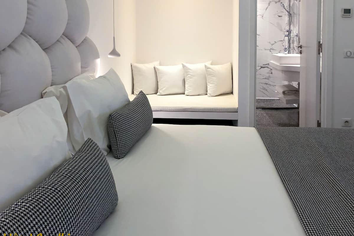 A modern hotel room with a large bed, white bedding, a sofa with white cushions, and a bathroom visible through an open door.