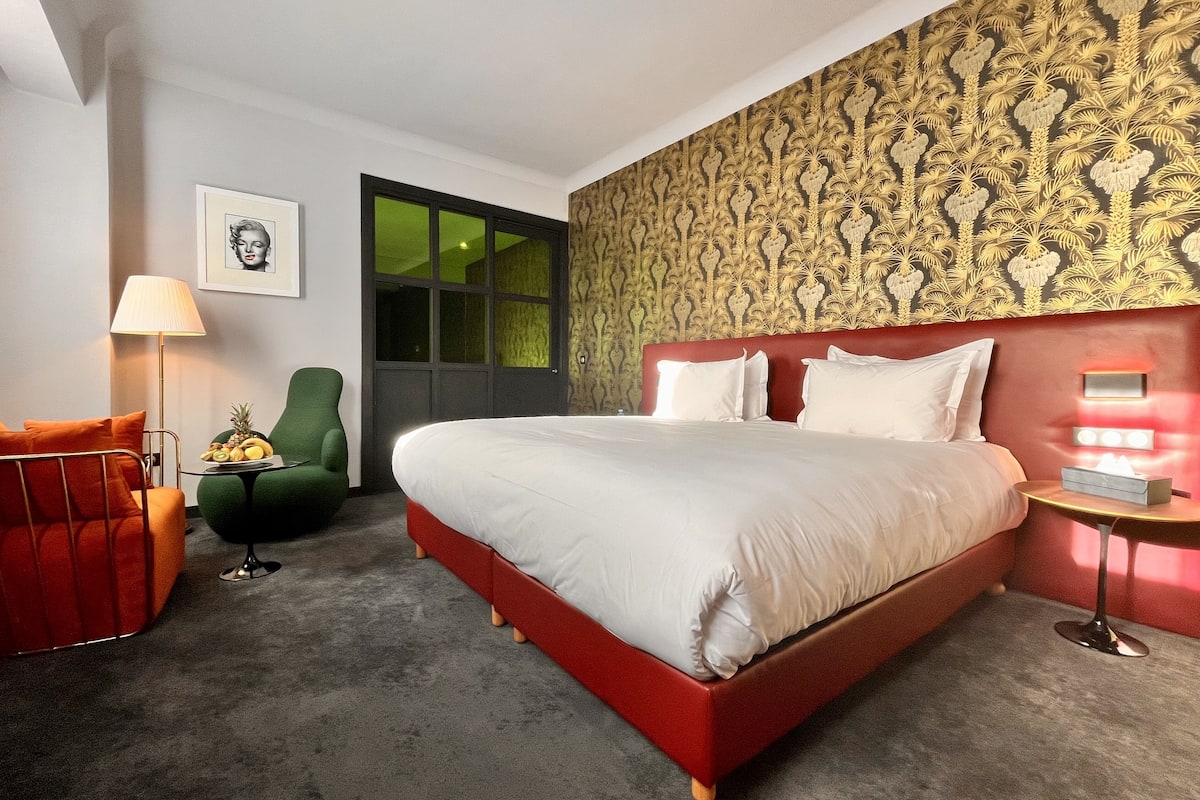 A hotel room with a red bed, a green armchair, a small table with fruit, a lamp, and a wall with a patterned design.