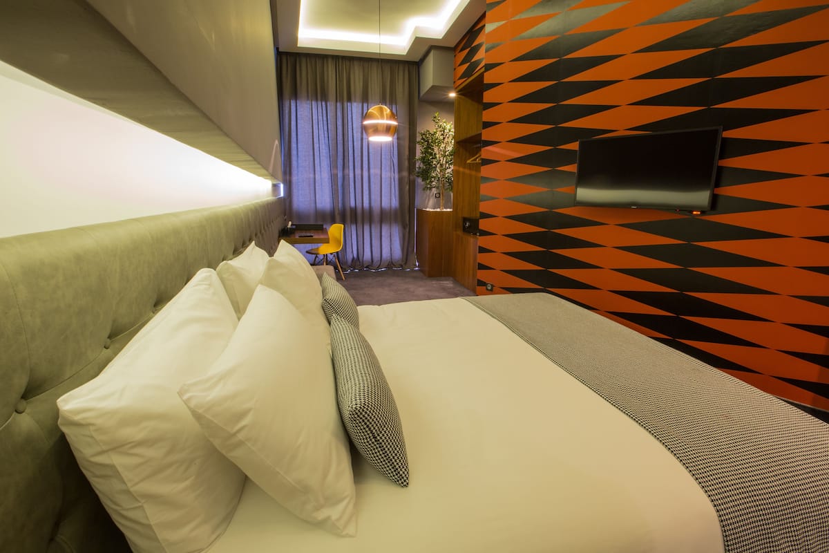 A hotel room with a bed, pillows, a television mounted on the wall, and a patterned wall.