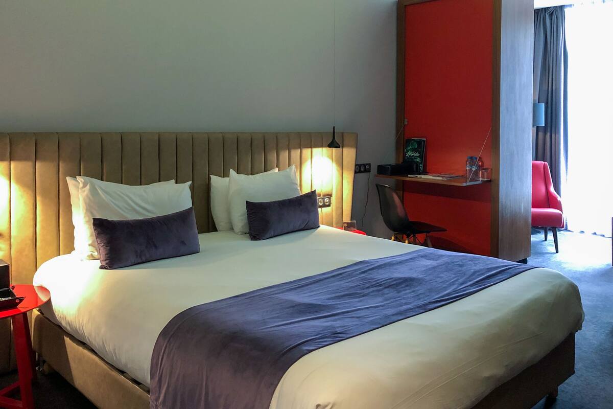A hotel room with a large bed, a red chair, a desk, and a red wall.