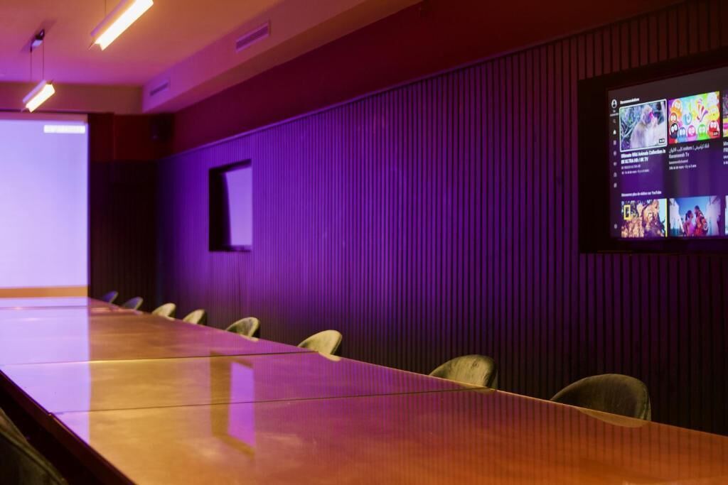 Une salle de conférence avec une longue table et des chaises, un écran de projection affichant du contenu et un mur éclairé en violet.