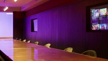 Une salle de conférence avec une longue table et des chaises, un écran de projection affichant du contenu et un mur éclairé en violet.
