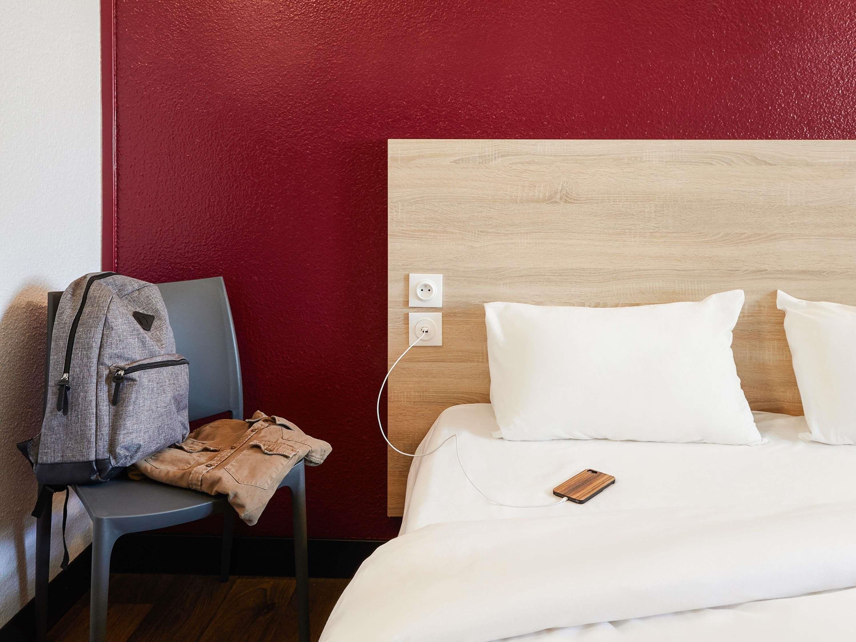 Una habitación de hotel con una cama, una silla, una mochila y un cabecero de madera.