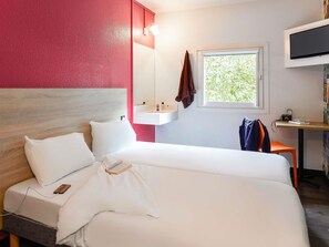 A hotel room with a bed, a bedside table with a lamp, a TV mounted on the wall, and a window with a view of greenery.